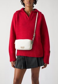 Maglione rosso lavorato a maglia con colletto, abbinato a una gonna plissettata grigia e a una borsa a tracolla bianca in pelle con dettagli del logo dorati.
