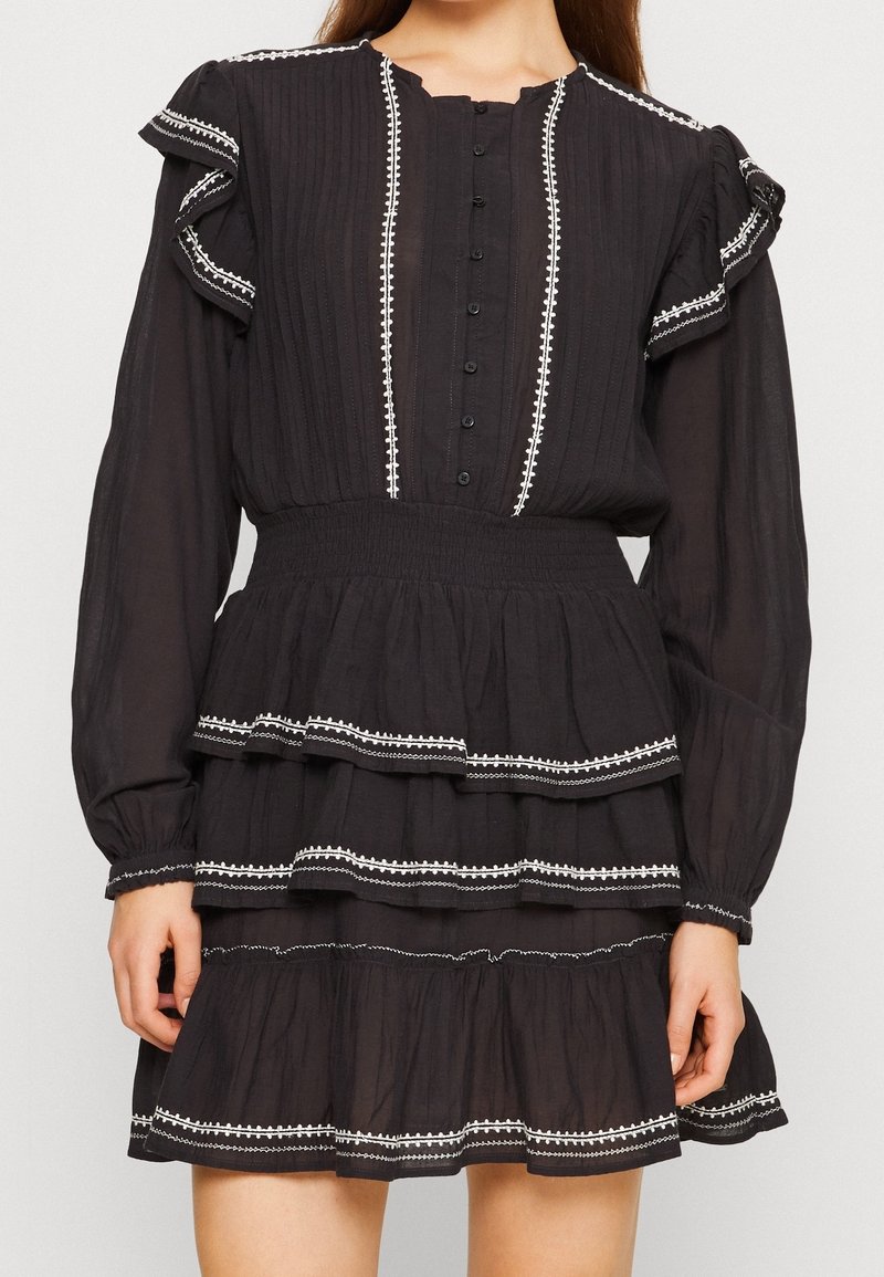 Robe noire à volants, à manches longues, avec des accents ondulés et des broderies blanches, dotée d'une fermeture à boutons à l'avant et d'une taille smockée.