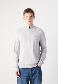 Lys grå zip-up genser, teksturert stoff, ribbet krage og mansjetter, med en liten svart logo på brystet, sammen med blå denimjeans.
