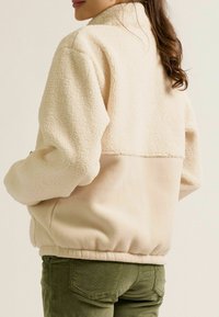 Cremfarbene Fleecejacke mit hohem Kragen, strukturiertem Oberkörper und glattem unteren Paneel; elastischer Bund; gemütliches und lässiges Design.