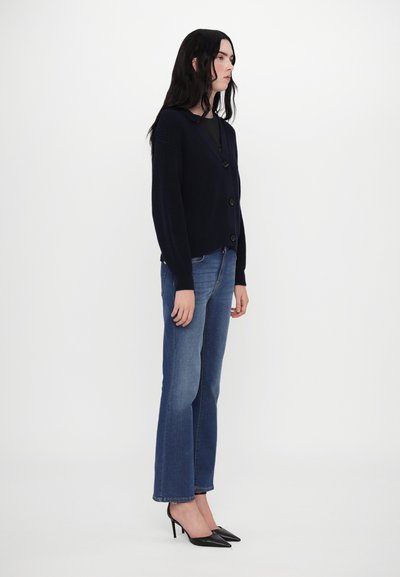 Marineblauer gerippter Cardigan mit V-Ausschnitt und Knopfverschluss, kombiniert mit hellblauen geraden Jeans und schwarzen spitzen Absatzschuhen.