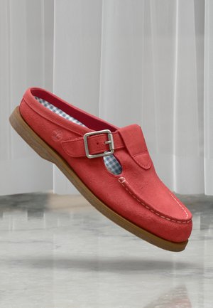 Mocassin en daim rouge avec boucle argentée, doublure intérieure à carreaux bleus, coutures blanches et semelle marron clair sur sol réfléchissant.