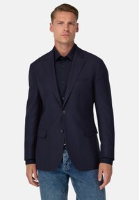 Marinblå blazer med texturerad finish, två framfickor, enkelknappslåsning och slagkragar, bärs över en mörk knapp-up skjorta.