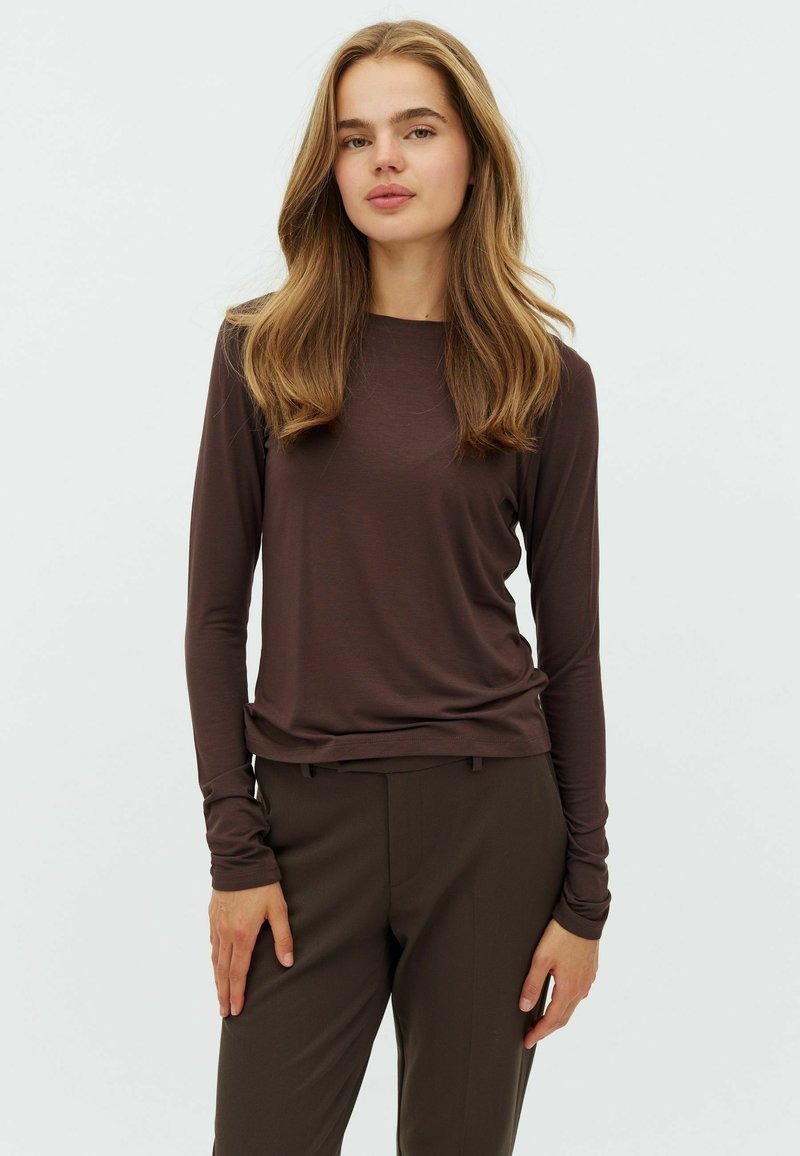 Jeune femme aux longs cheveux châtain clair portant un haut marron ajusté à manches longues et un pantalon marron foncé, debout devant un fond uni.