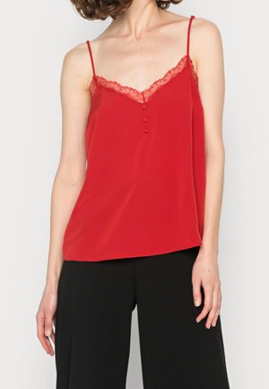 Femme portant un caraco rouge orné de dentelle avec de fines bretelles et un pantalon noir à jambes larges, debout devant un fond uni.