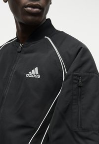 Μαύρο τζάκετ Adidas με φερμουάρ, με λευκή διακόσμηση και λογότυπο, που διαθέτει τσέπη με φερμουάρ στο επάνω μέρος του μανικιού, φορεμένο από ένα άτομο.