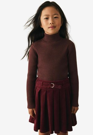 Marlburgunder Rollkragenpullover mit gerippter Textur, kombiniert mit einem plissierten Cordrock in passender Farbe und einem Gürtel mit silberner runder Schnalle.