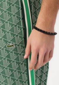 Zelená látka s geometrickým vzorem, obsahující bílý zip a zelené pruhované akcenty. Na zápěstí je nošena černá řetízková náramek.