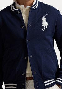 Marinblå varsityjacka med vit randig krage och manschetter, med en vit broderad polo-spelaren logotyp på vänster bröst.