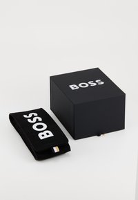 Juodas dėžutė su balta užrašu "BOSS" viršuje ir atitinkama juoda krepšys, kuriame užrašytas "BOSS" paryškintai. Krepšys turi mažą auksinės spalvos etiketę.