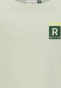 Jasnoszara koszulka z krótkim rękawem z małym ciemnozielonym kwadratowym logo i żółtym napisem „The Orig” na górnej prawej części klatki piersiowej.