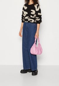 Frau trägt einen schwarz-beige gemusterten Pullover, weit geschnittene blaue Jeans, schwarze grobe Stiefel und h�ält eine rosa geraffte Handtasche an ihrer Seite.