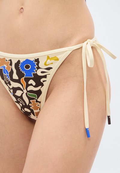 Κοντινό πλάνο ενός ατόμου που φοράει μπεζ μαγιό με πολύχρωμα αφηρημένα σχέδια και δεσίματα στο πλάι, που δείχνει μέρος της κάτω κοιλιάς και του μηρού.