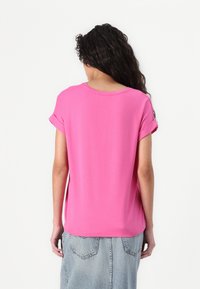 Femme aux longs cheveux bouclés portant un t-shirt manches courtes rose vif et une jupe en jean bleu clair, vue de dos sur un fond blanc uni.