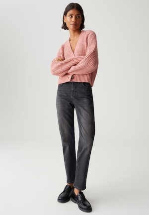 Femme portant un cardigan en tricot épais rose, un jean gris foncé et des chaussures noires à lacets, se tenant les bras croisés sur un fond uni.