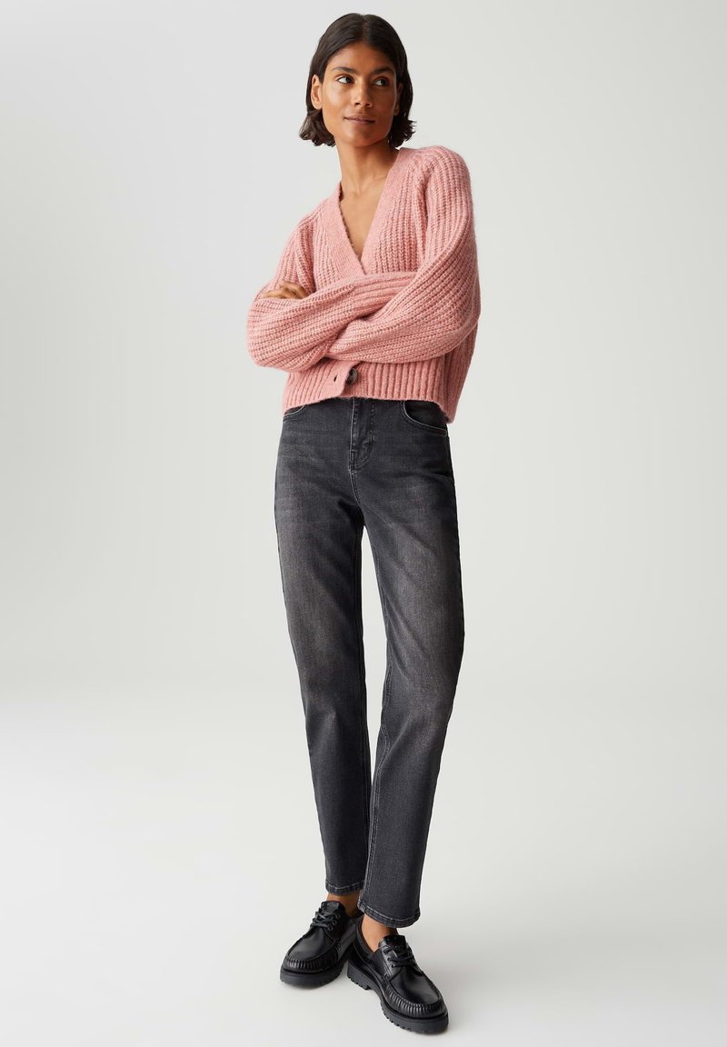 Donna che indossa un cardigan spesso rosa, jeans grigio scuro e scarpe nere con stringhe, in piedi con le braccia incrociate su uno sfondo neutro.