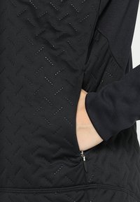 Main dans la poche zippée d'une veste noire texturée avec de petits trous de ventilation et des manches longues.