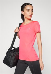 Tett, kortermet, ribbestrikket rosa treningstrøye med blå logo, kombinert med en svart treningsbag med nettingdetaljer og skulderrem.