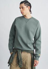 Pilko žalios spalvos džemperis su ribotu apvaliu kaklu ir raglano rankovėmis. Derinamas su smėlio spalvos kelnių su kišenėmis modeliu ir žalia kamufliažine medžiaga, surišta aplink juosmenį.