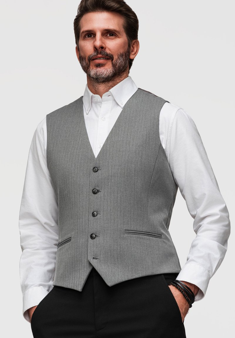 Homme barbu portant un gilet gris boutonné sur une chemise blanche et un pantalon noir, les mains dans les poches, sur un fond neutre.