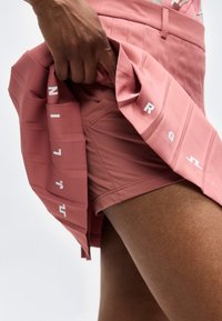 Χέρι σηκώνει ροζ πλισέ φούστα αποκαλύπτοντας ταιριαστά εφαρμοστά σορτς από κάτω με λευκά τυπωμένα γράμματα στα πτυχώματα της φούστας.