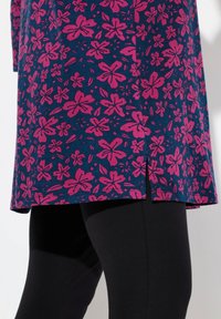 Modrá tunika s růžovým květinovým vzorem, s rovnými lemy a bočním rozparkem, spárovaná s černými legínami pro lepší kontrast.