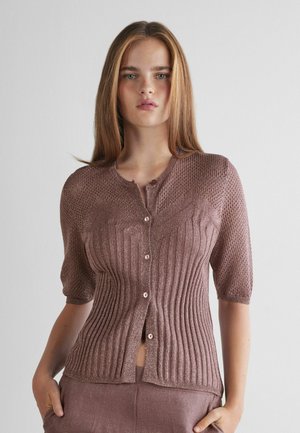 Junge Frau mit glatten hellbraunen Haaren trägt ein mauvefarbenes, kurzärmeliges, gestricktes Oberteil mit Knöpfen und passende Hosen, stehend vor einem schlichten weißen Hintergrund.