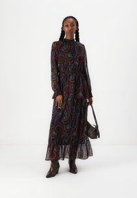 Vestido longo escuro de padrão paisley com bainha de franja, cintura ajustada, decote alto e mangas longas. Usado com botas de salto castanhas e uma bolsa pequena.