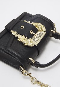 Borsa a tracolla in pelle nera texturizzata con fibbia dorata ornamentale e etichetta "Versace Jeans Couture", dotata di un manico in pelle e una tracolla in catena dorata.