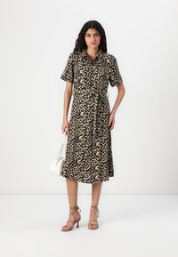 Klänning med knäppning i beige med svart leopardmönster, korta ärmar, midja med bälte och midi-längd. Bärs med sandfärgade heels med remmar och en vit väska.