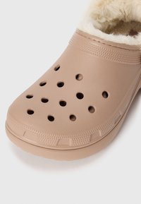 Zoccoli beige con una superficie sintetica liscia, dotati di fori di ventilazione, un rivestimento morbido e una suola texturizzata per una presa migliore.