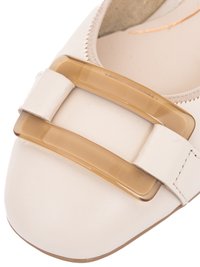 Nahaufnahme eines flachen Schuhs in Cremefarbe mit großer, durchscheinender beiger rechteckiger Schnalle an der Spitze.