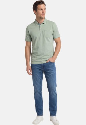 Mann trägt ein hellgrünes Poloshirt mit kurzen Ärmeln, blaue Jeans und weiße Turnschuhe, steht mit einer Hand in der Tasche und schaut zur Seite.