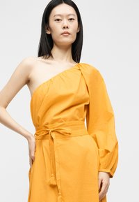 Femme aux cheveux noirs et raides portant une robe orange vif à une épaule avec une longue manche bouffante et une ceinture nouée à la taille.