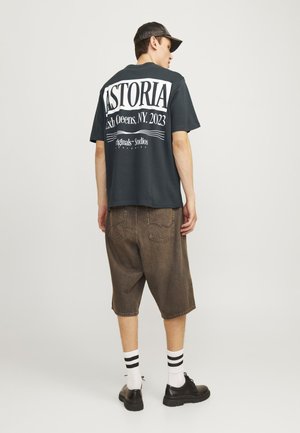 T-shirt verde oscuro con impresión blanca en la parte de atrás; combinado con pantalones cortos marrones hasta la rodilla y zapatos negros. Los calcetines blancos tienen rayas negras.