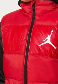 Czerwona kamizelka puchowa z wysokim kołnierzem i czarnymi akcentami. Posiada srebrny zamek błyskawiczny oraz biały logo Jordan nadrukowane na piersiach.
