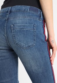 Jeans de mezclilla en azul claro con un acabado desgastado, con un bolsillo trasero, detalle de franja roja en el lateral y una textura suave.