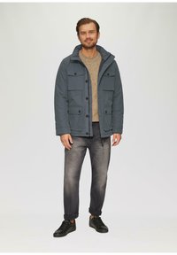 Männliche blaue Kapuzenjacke mit mehreren Fronttaschen, kombiniert mit einem beigen Pullover und dunklen Jeans, sowie schwarzen Sneakern.