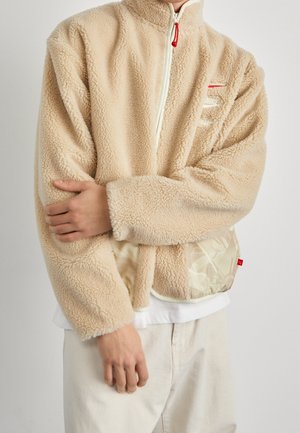 Person iført beige shearling fleecejakke med hvid lynlås og logo, cremefarvede bukser og holder modsatte arm på tværs af kroppen.