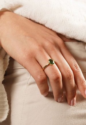 Goldring mit einem ovalen grünen Edelstein, getragen an einer Hand mit heller Haut, vor einem beigen Stoffhintergrund. Glatte Textur und einfaches Design.
