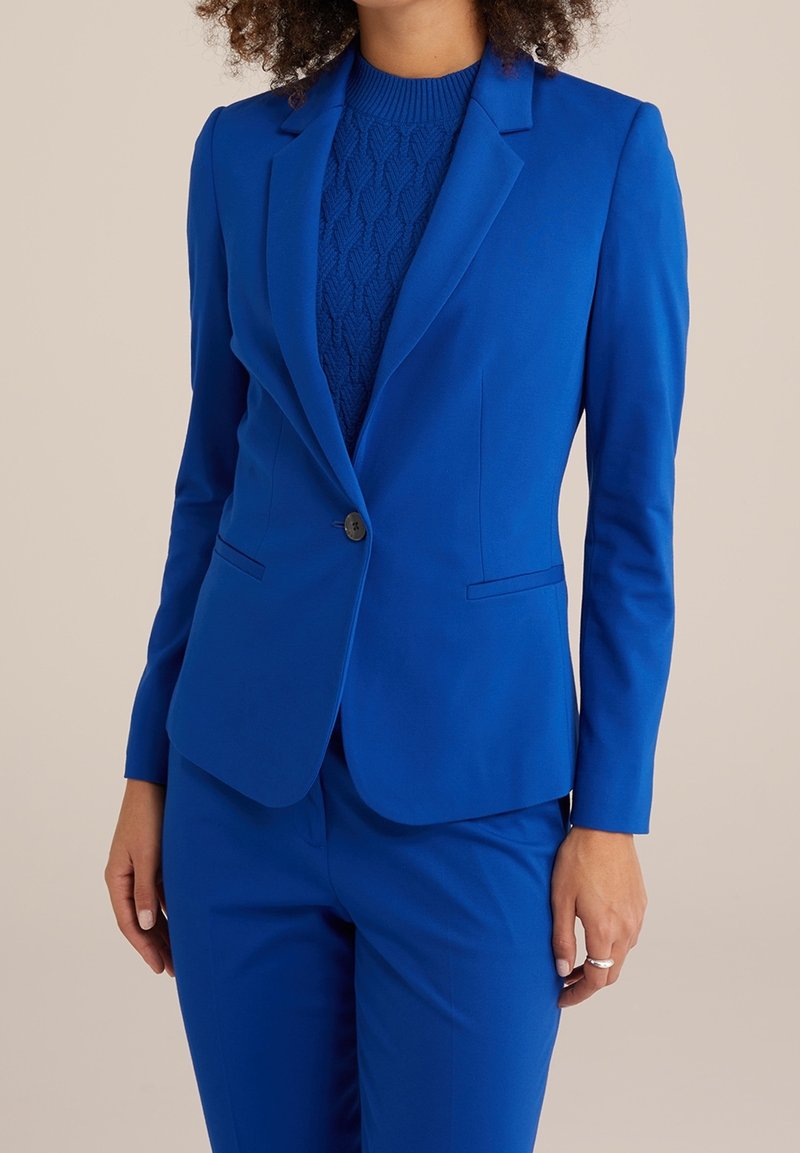 Blazer blu su misura con rever a contratto, chiusura con un solo bottone e due tasche laterali. Indossato sopra un top in maglia blu a trama. Tessuto liscio.