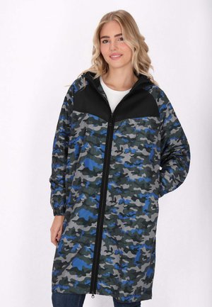 Jonge vrouw met blond haar die een lange, met rits sluitende jas draagt in blauw, grijs en zwart camouflagepatroon, met capuchon, staand tegen een effen lichte achtergrond.