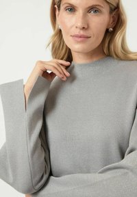 Szary, fakturowany top z wysokim dekoltem i szerokimi, rozciętymi rękawami, z delikatnym połyskiem. Ręka modelki spoczywa w pobliżu dekoltu.