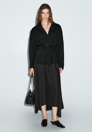 Maglione avvolgente nero in maglia abbinato a una gonna lunga nera, completato da scarpe slip-on nere e una borsa rigida. Sfondo neutro.