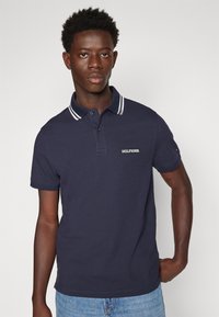 Polo shirt navy con colletto a strisce bianche, maniche corte e logo ricamato sul petto. Il tessuto appare liscio e il design è casual.