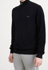 Zwarte gebreide trui met een hoge kraag, een gestructureerd breipatroon en een klein logo op de borst, gecombineerd met lichtgrijze broek.