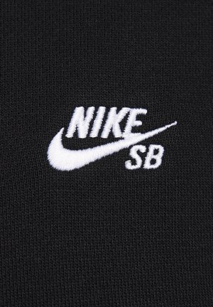 Melnas auduma ar teksturētu adījumu, ar baltu izšūtu "NIKE SB" logotipu un swoosh, kas demonstrē krāsu un zīmola kontrastu.
