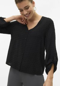 Blusa nera con scollo a V, maniche lunghe e dettagli con bottoni. Tessuto testurizzato con punti in rilievo. Materiale morbido e leggero.