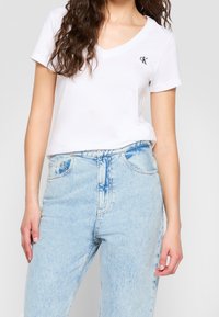 Bílý bavlněný T-shirt s výstřihem do V a černým logem "CK", spárovaný se světle modrými džínami s vysokým pasem, které mají klasický pětipedlový design.