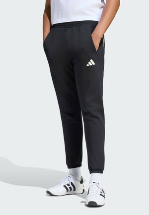 Osoba nosząca czarne spodnie dresowe Adidas z białym logo, białe skarpetki i białe buty sportowe z czarnymi paskami na jasnym tle.
